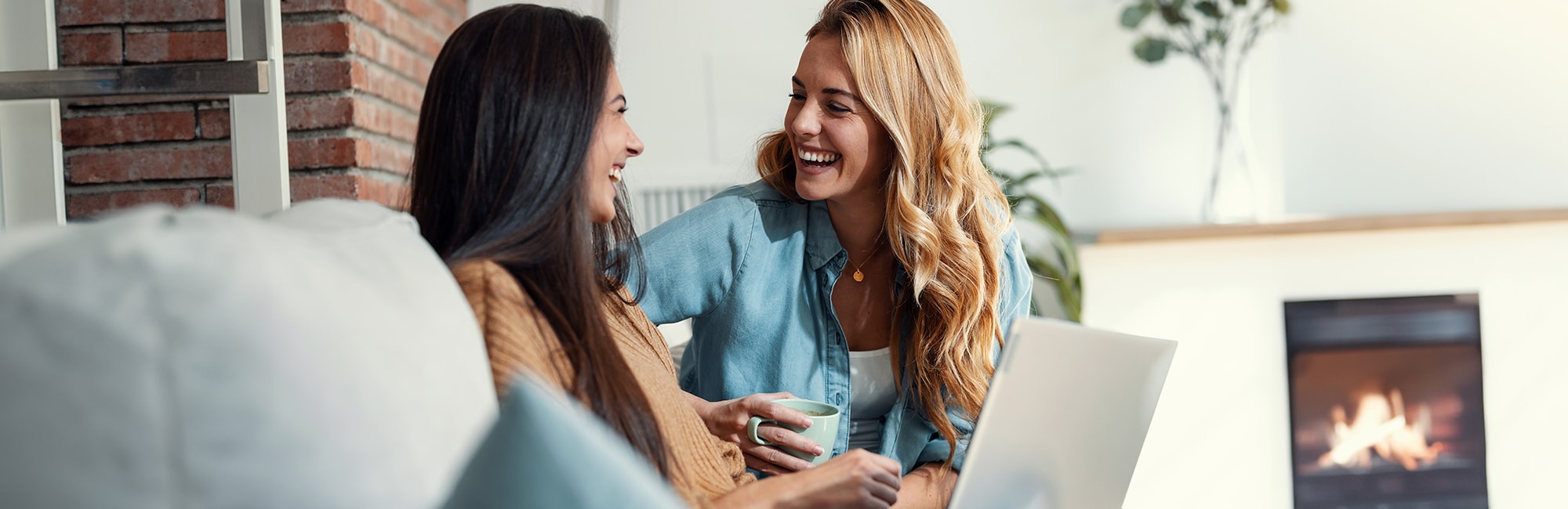 Two women are sitting on a couch, laughing together. One of them holds a mug, whilst the other has a laptop on her lap.