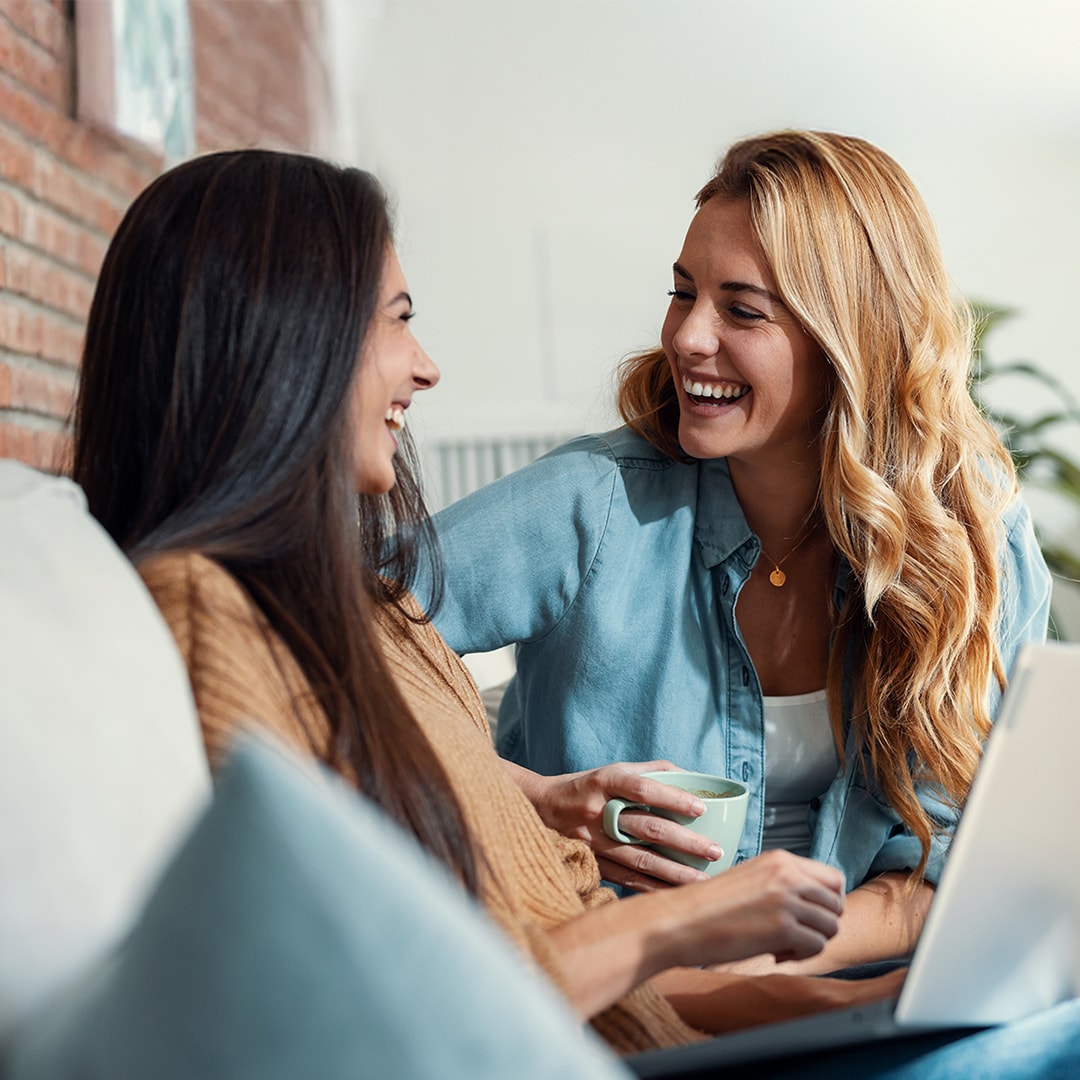 Two women are sitting on a couch, laughing together. One of them holds a mug, whilst the other has a laptop on her lap. 